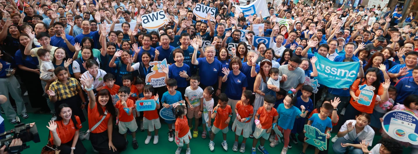 Large crowd waving, holding signs with 'AWS' and 'SSMC' logos. People wear blue and orange shirts.